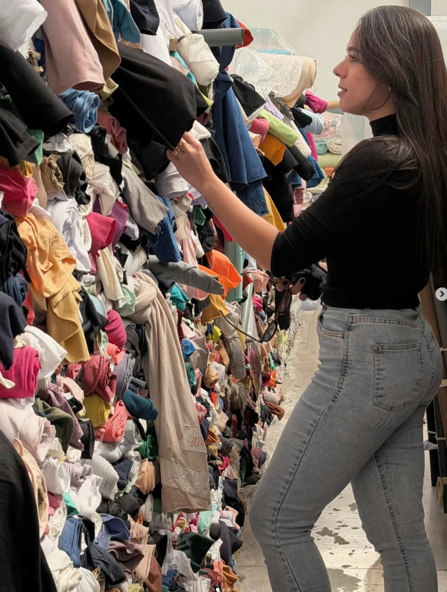 Thaina escolhendo tecidos em meio a pilhas de malhas coloridas
